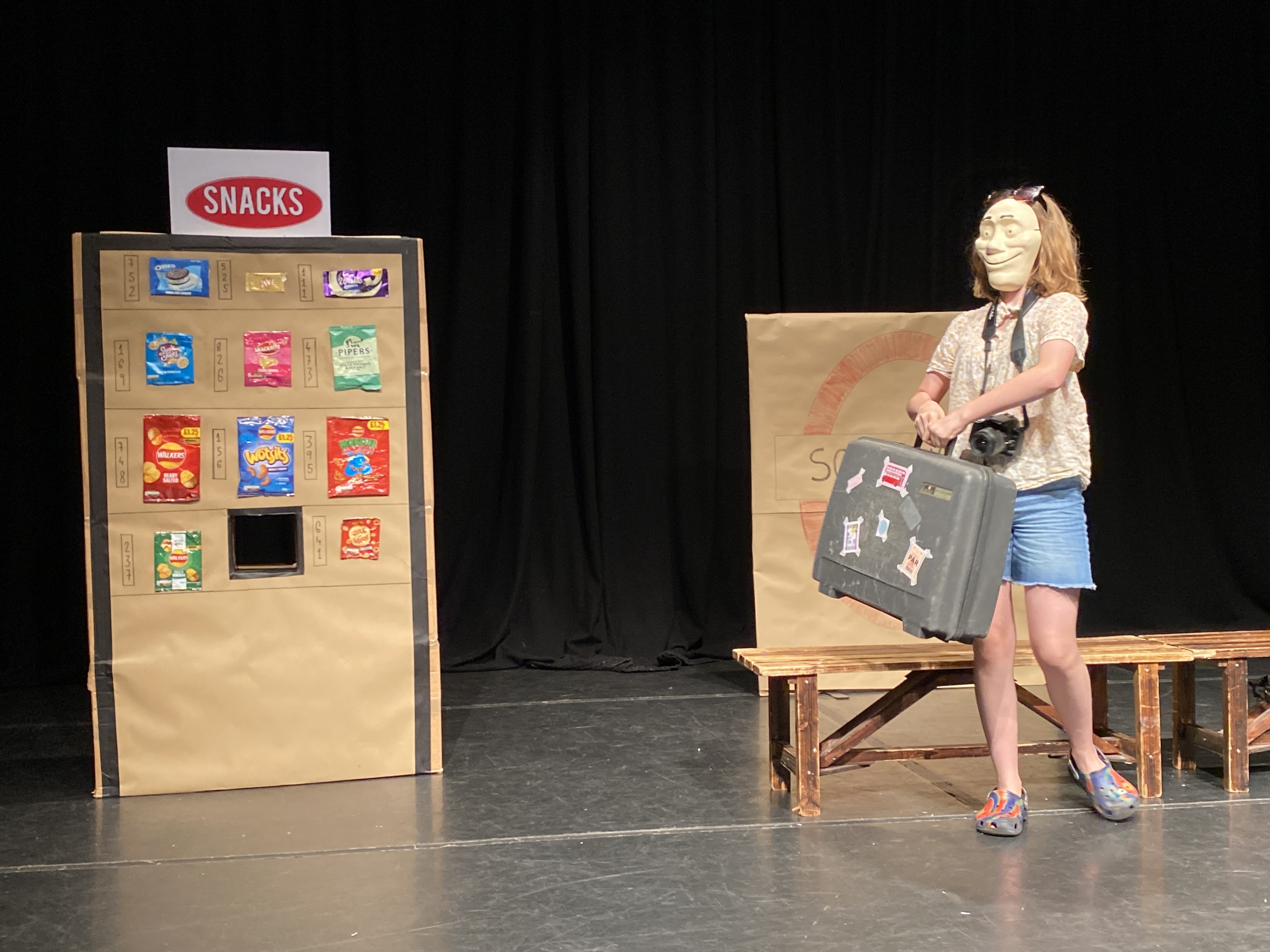 A young woman wearing a Trestle Intermediate Mask is standing next to a booth filled with snacks. The scene is indoors, featuring various pieces of furniture including a chair. A young woman wearing a Trestle Intermediate Mask is standing next to a booth filled with snacks. The scene is indoors, featuring various pieces of furniture including a chair.